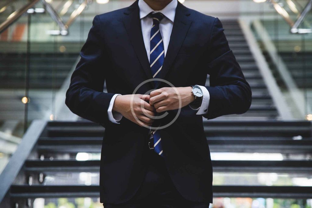 A person in a suit adjusts their jacket sleeves while standing in front of a staircase.