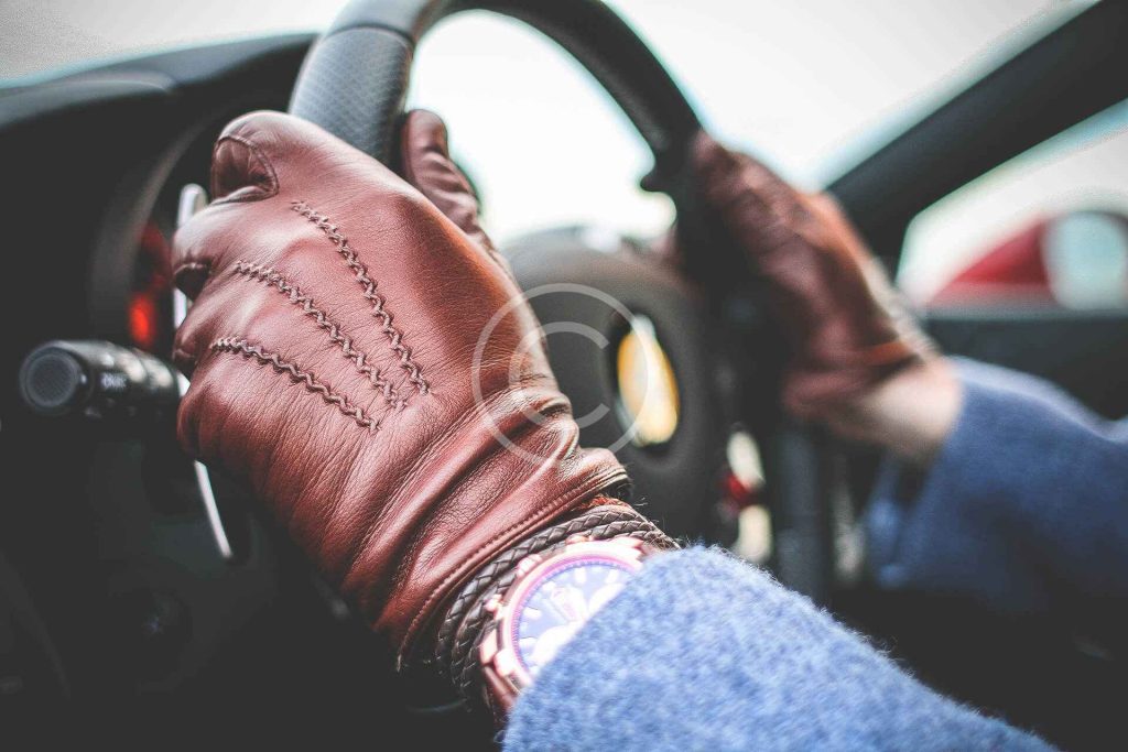 A person wearing brown leather gloves and a wristwatch is gripping a car steering wheel, with part of the dashboard visible.