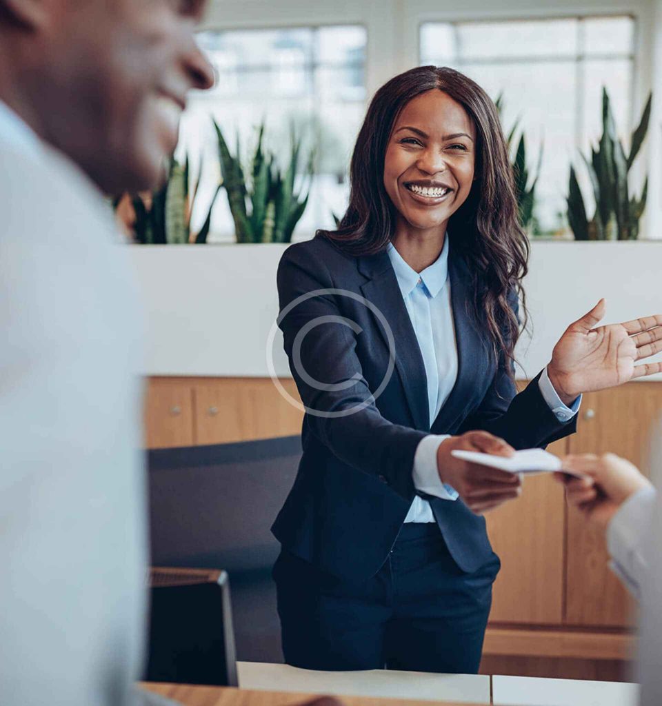 A woman in business attire smiles as she hands a document to someone across a desk in a modern office setting.