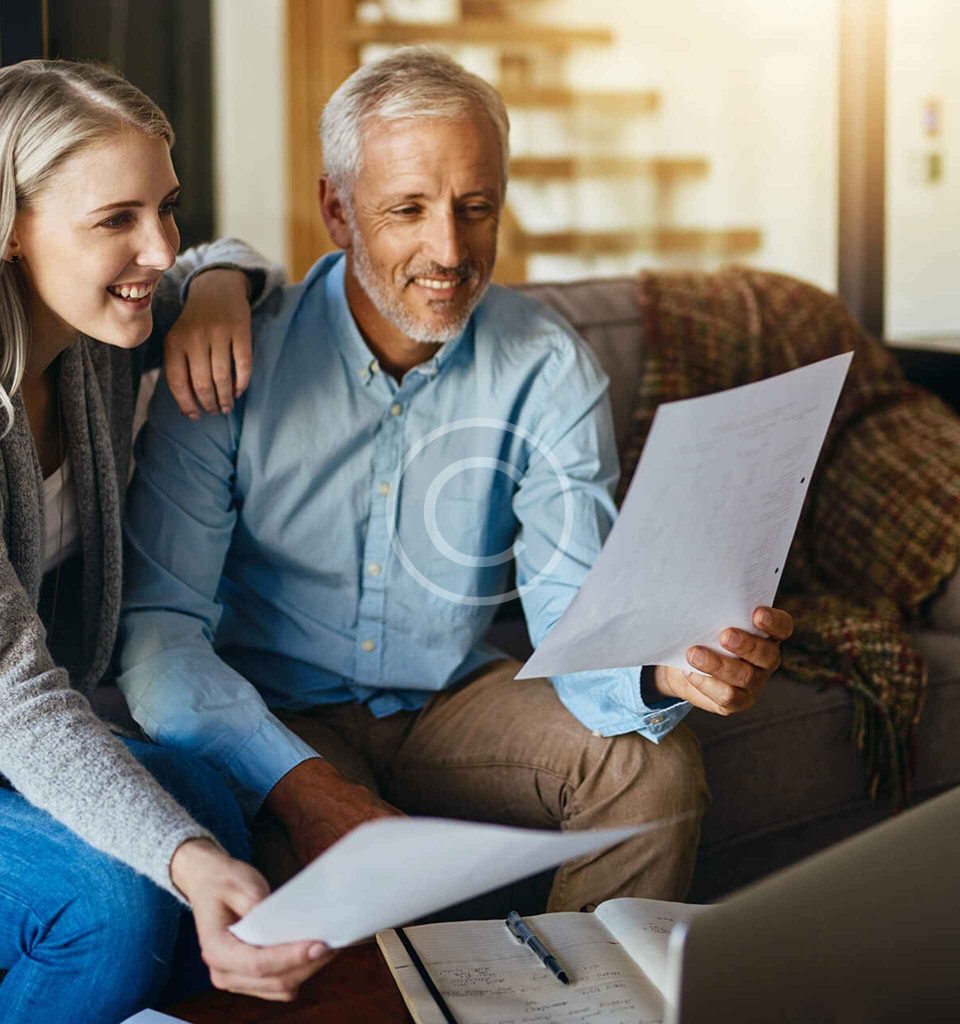 An older man and a younger woman sit on a couch, smiling while reviewing paperwork together, with a notebook and pen on the table in front of them.