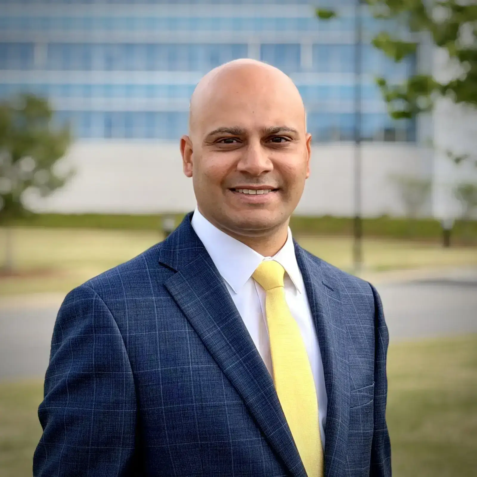 A criminal defense attorney in Lawrenceville, GA, wearing a blue suit and yellow tie, stands outdoors in front of a modern building, smiling at the camera.